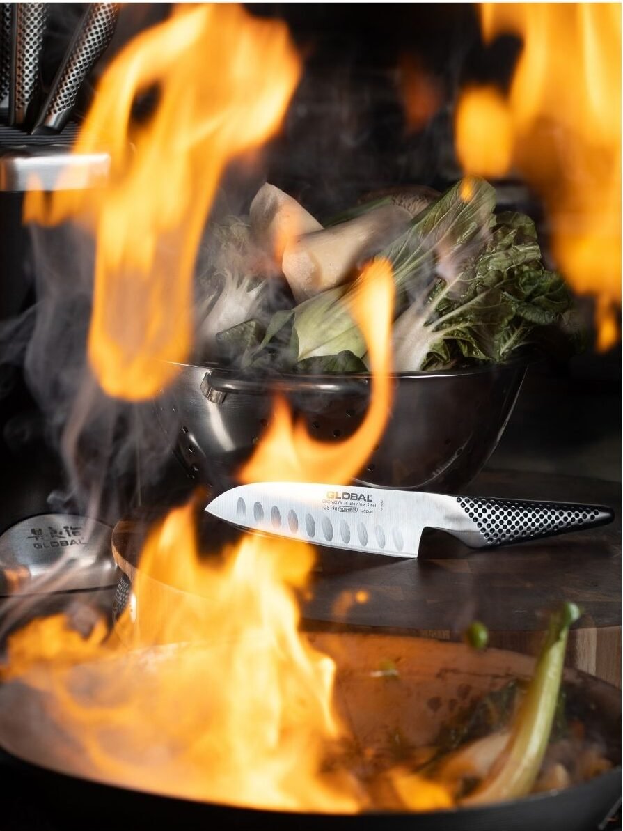 Global santoku knife on a board beside a wok in full flame, pak choi waiting in a steel colander
