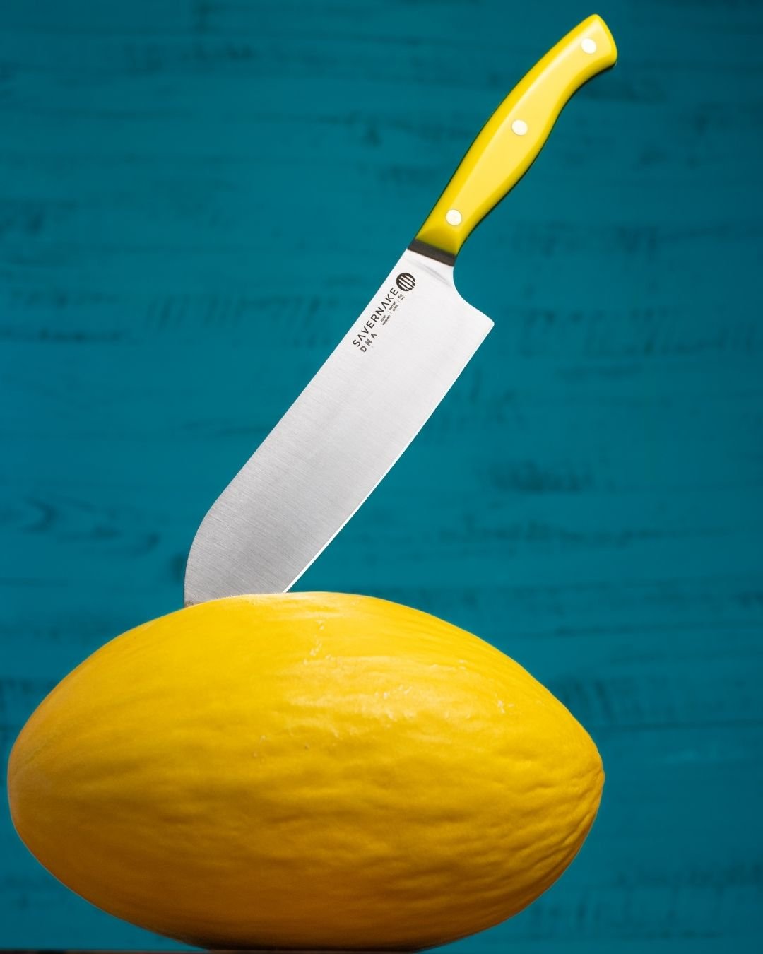 Savernake DNA chef's knife with a yellow handle cutting into a yellow melon on a teal background