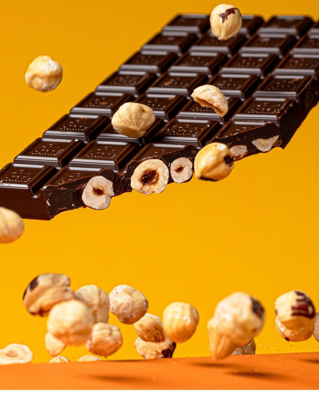 Dark chocolate bar with roasted hazelnuts falling around it against a yellow background