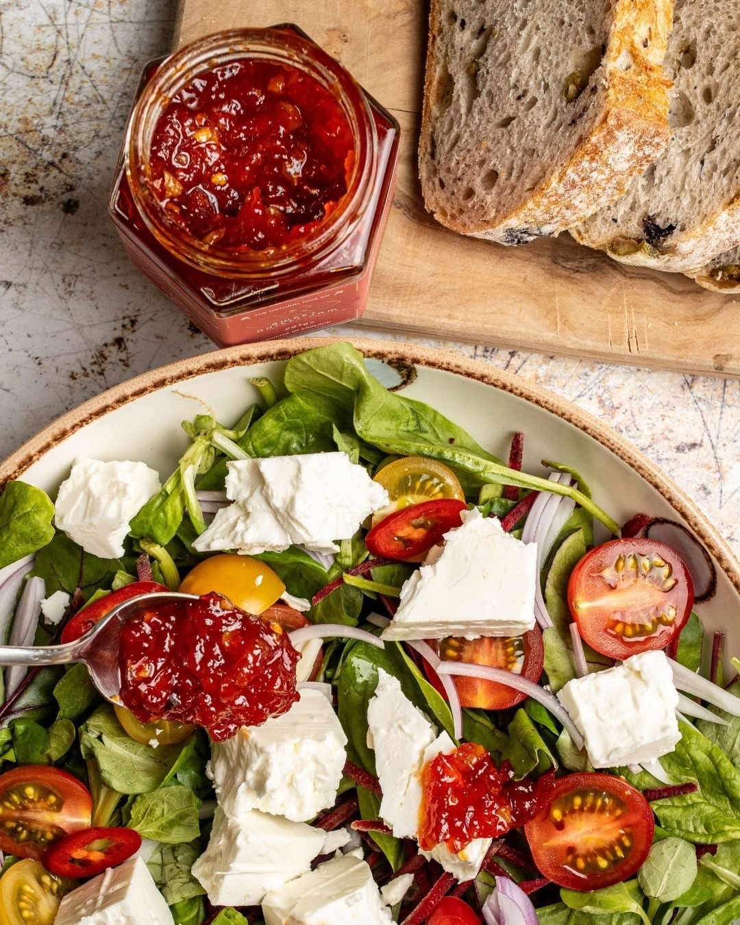 Overhead of a soft cheese and tomato salad with a jar of chilli jam and sliced sourdough