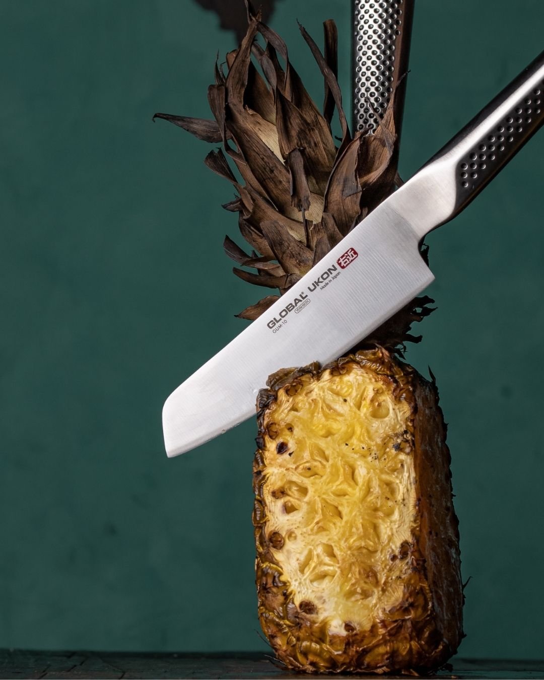 Global Ukon santoku resting on a halved pineapple against a dark green background