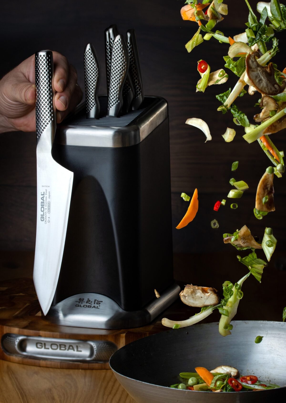 Hand lifting a Global chef's knife from its block, stir fry vegetables flying above a wok