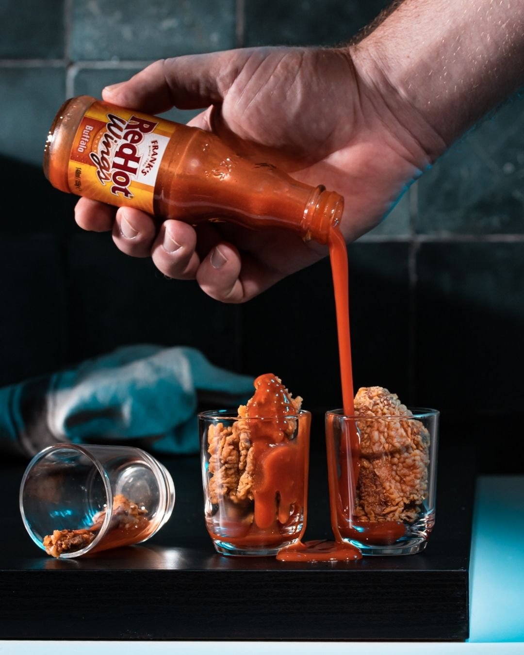 Hot sauce poured over fried chicken pieces served in glass tumblers on a dark counter