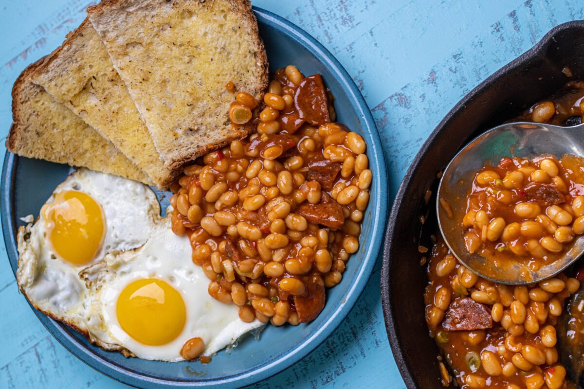 Overhead of baked beans with chorizo, fried eggs and buttered toast, cast iron pan alongside