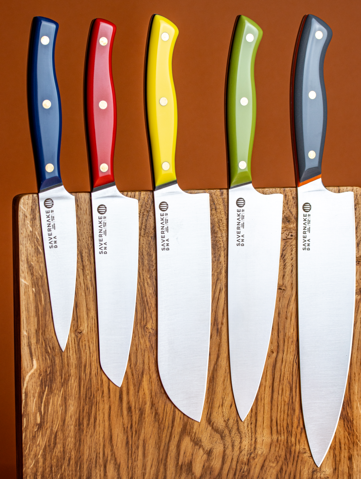 Five Savernake DNA chef's knives with coloured handles laid across an oak board