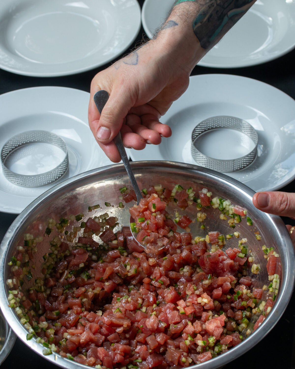 Chef spooning tuna tartare from a steel bowl into ring moulds, plates lined up ready