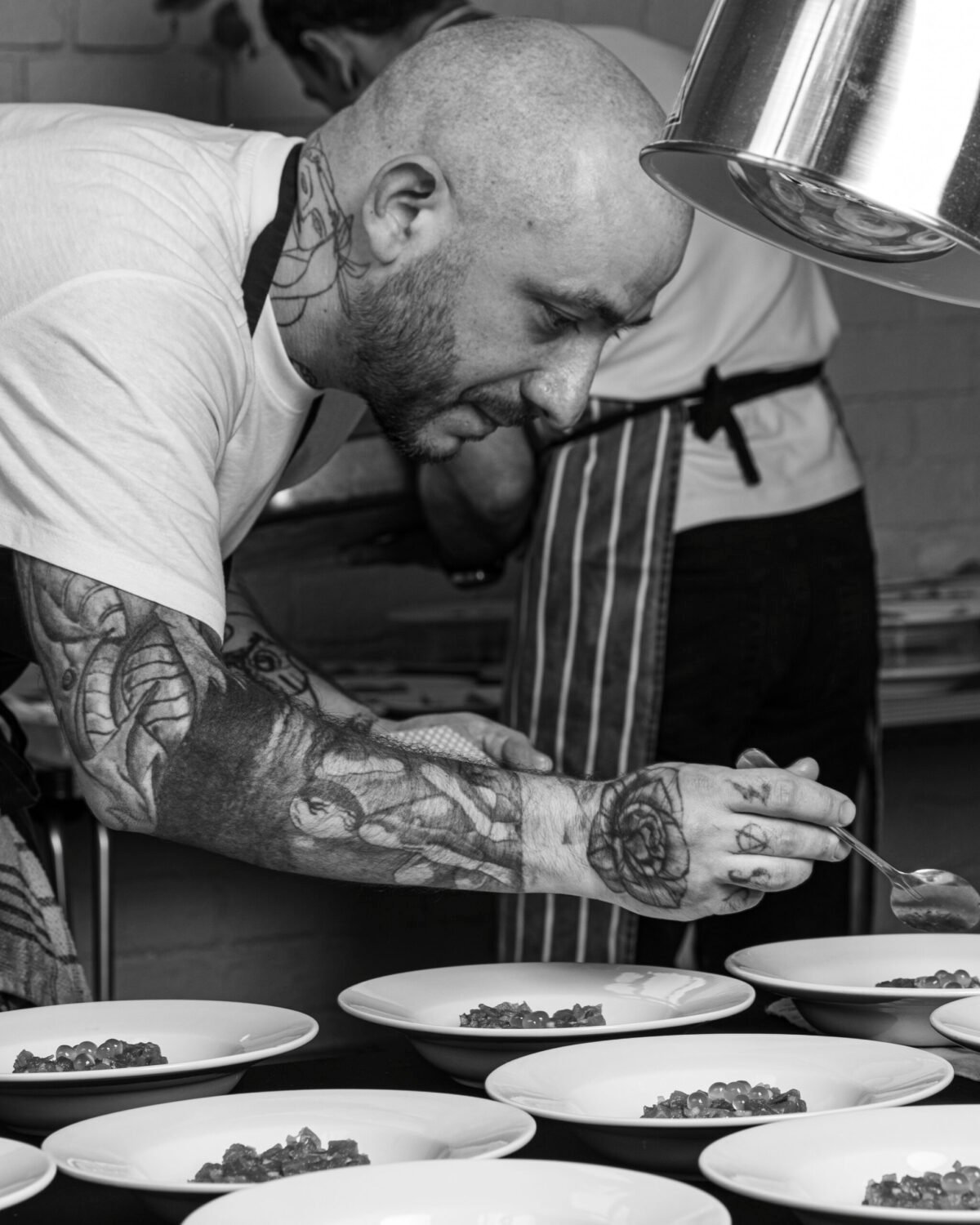 Black and white shot of a chef plating a line of dishes under a heat lamp