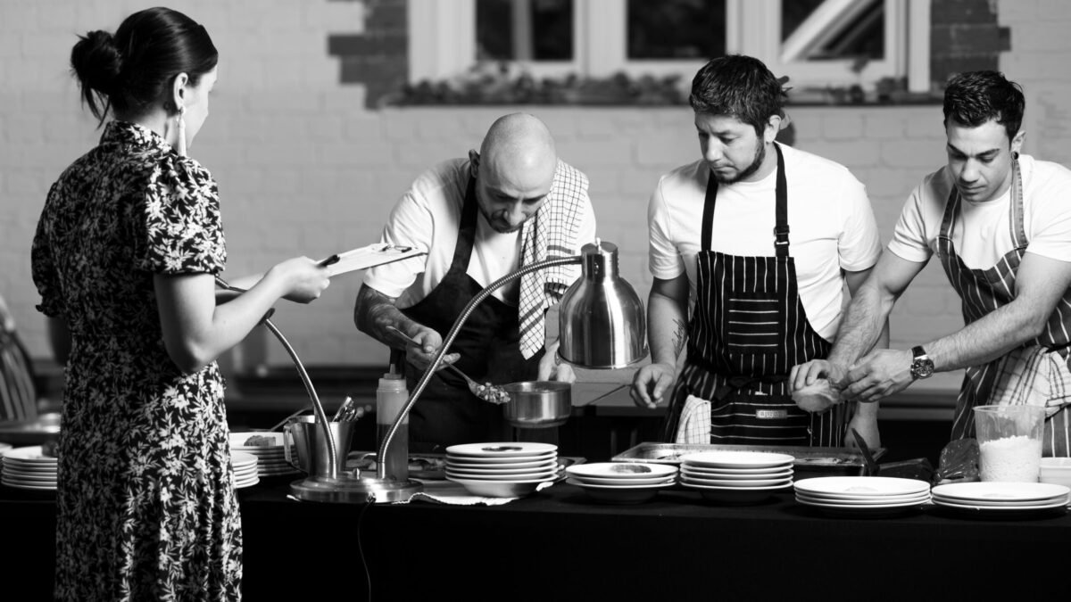 Black and white shot of four chefs working a catering pass, plates stacked ready