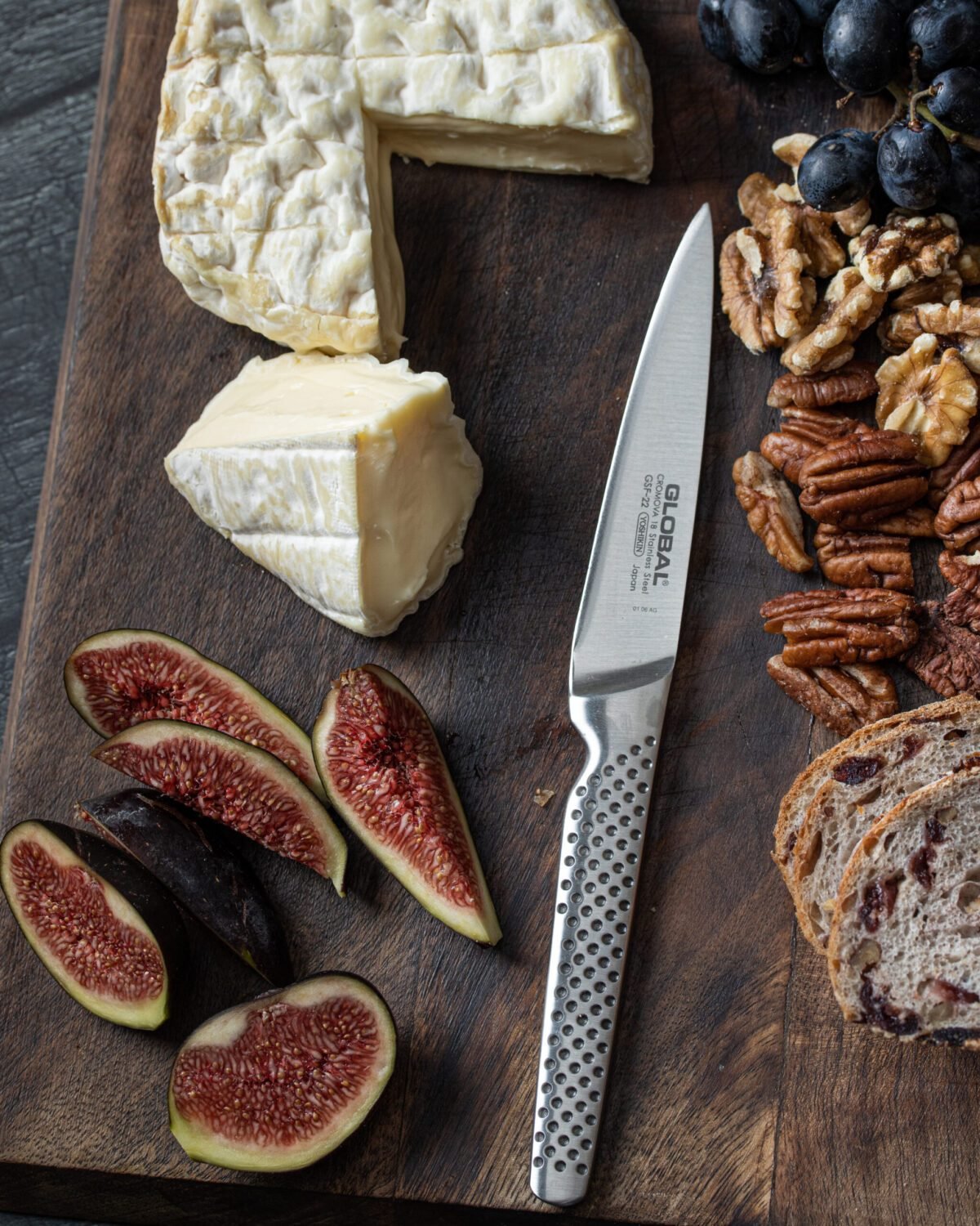 Brie, figs, grapes, walnuts and pecans on a dark board with a Global paring knife