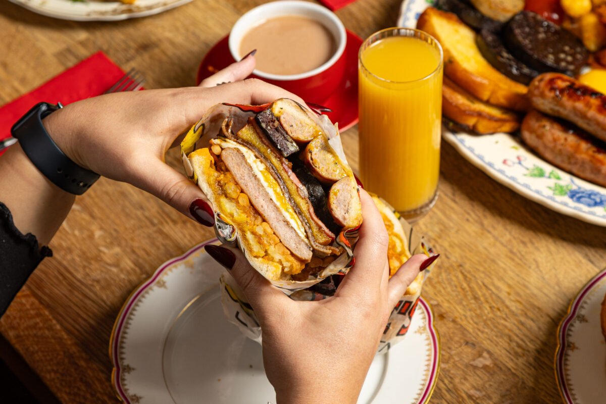 Hands holding a loaded breakfast sandwich over a plate, coffee and orange juice behind