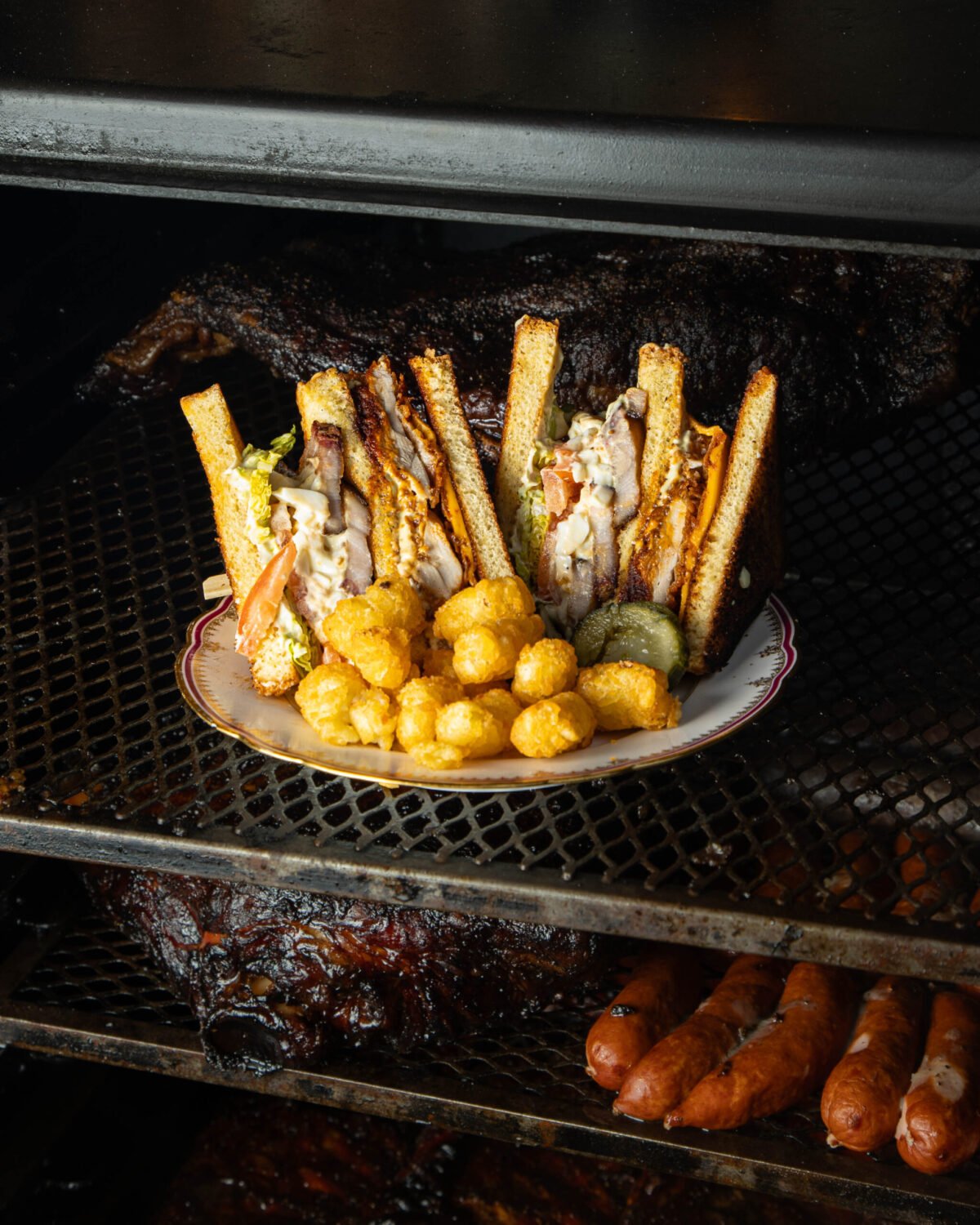 Club sandwich and tater tots plated inside a smoker, ribs and sausages on the racks below