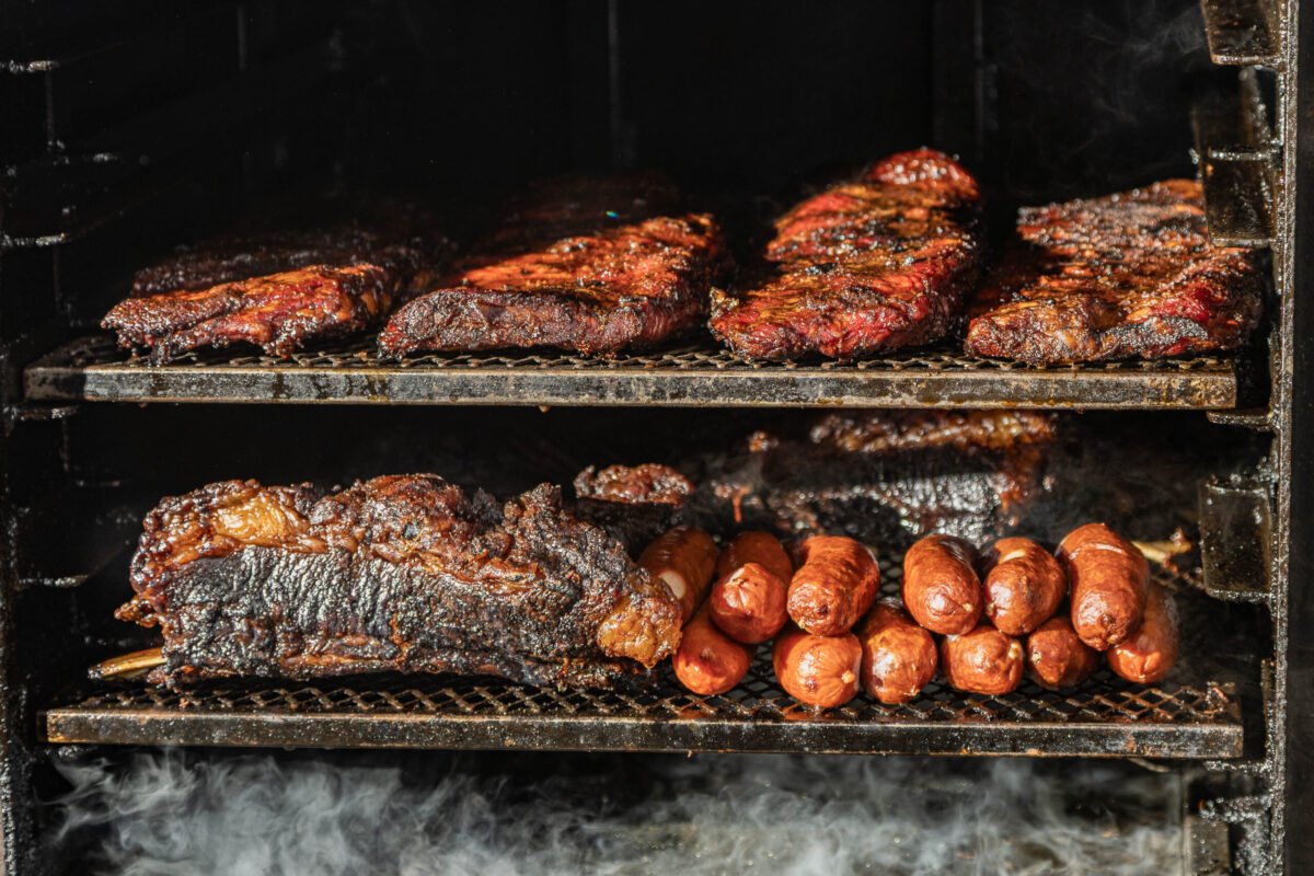 Racks of ribs, brisket and sausages inside a smoker with smoke drifting across the shelves
