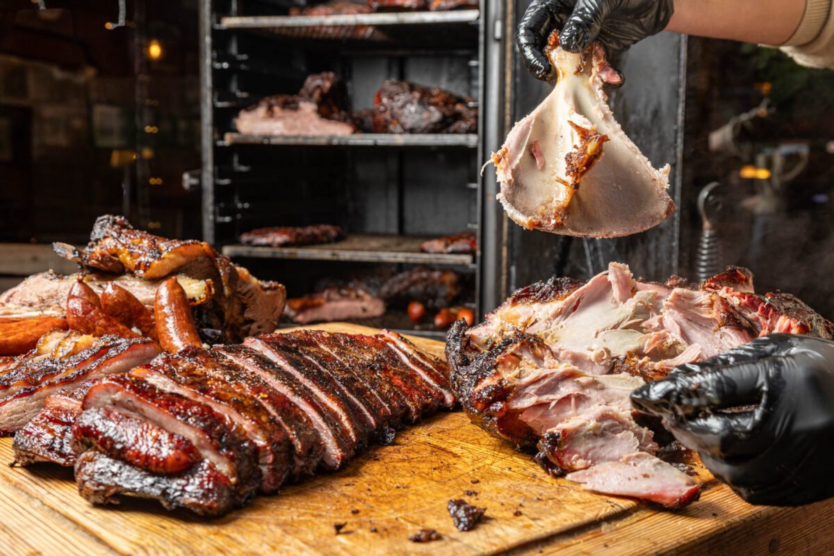 Gloved hands lifting smoked meat above a board of sliced brisket and ham