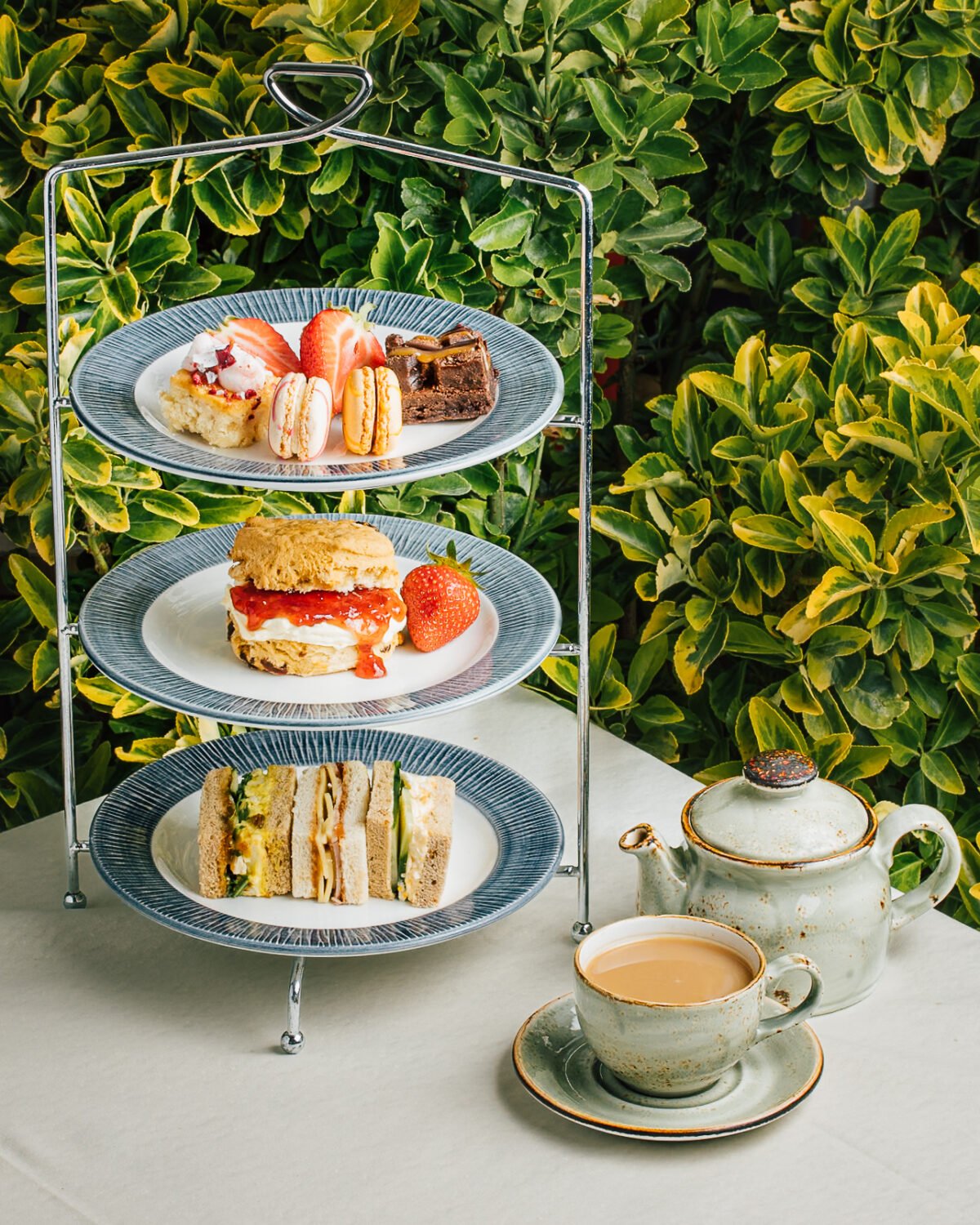 Three tier afternoon tea stand set outdoors against foliage with a teapot and cup of tea