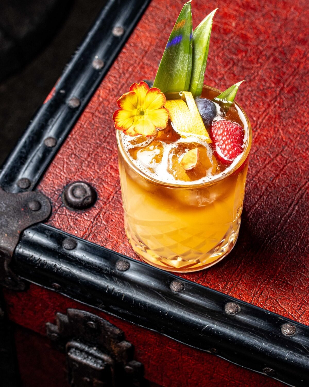 Amber cocktail garnished with pineapple leaf, raspberry and an edible flower on red leather