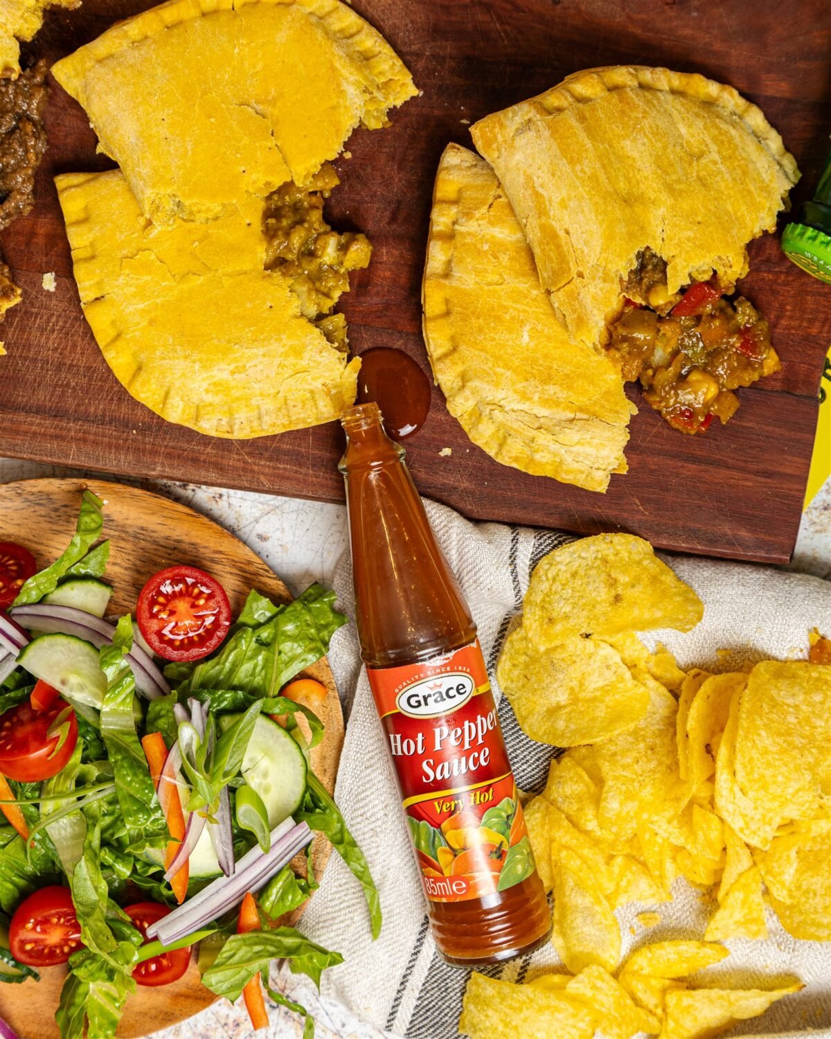 Two Jamaican patties on a dark board with a side salad and a bottle of pepper sauce