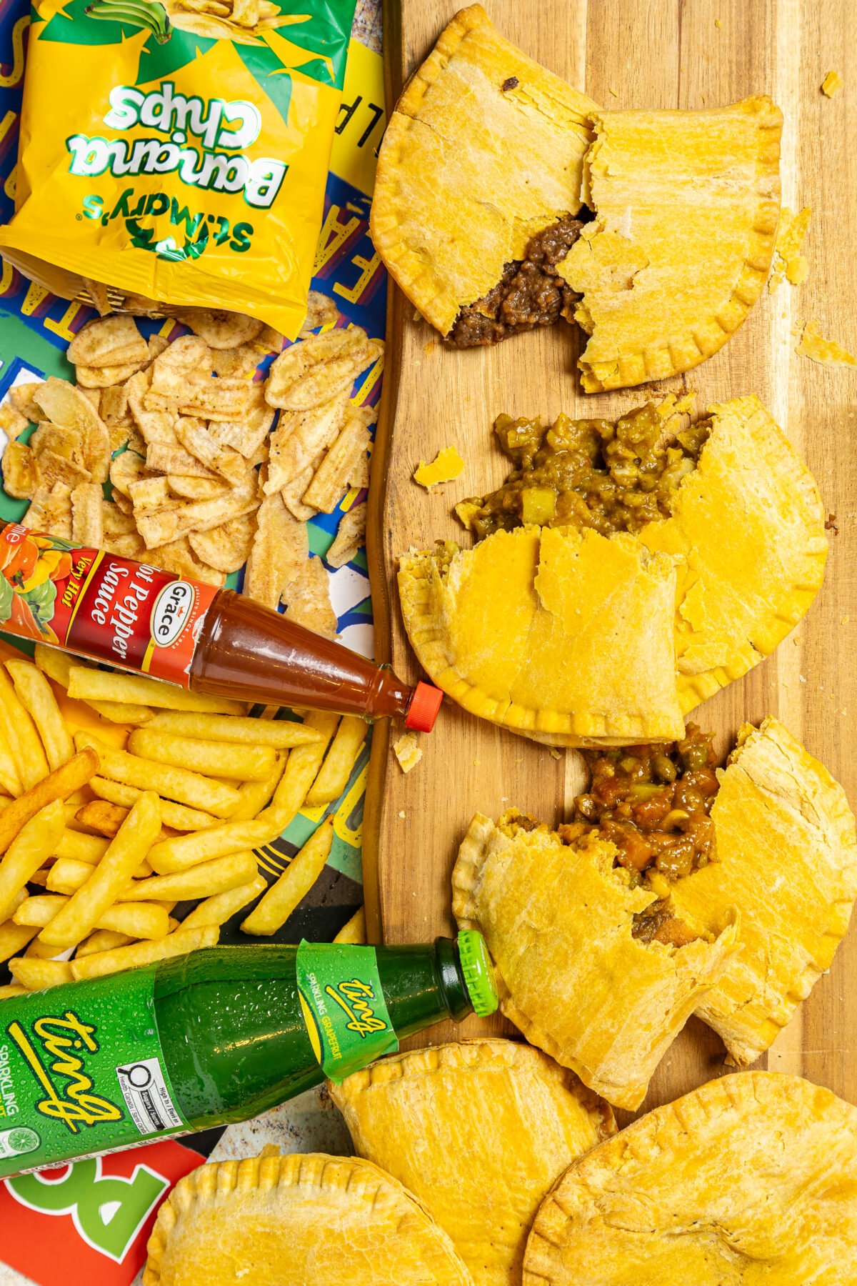 Overhead flatlay of Jamaican patties broken open on a board with banana chips, pepper sauce and fries