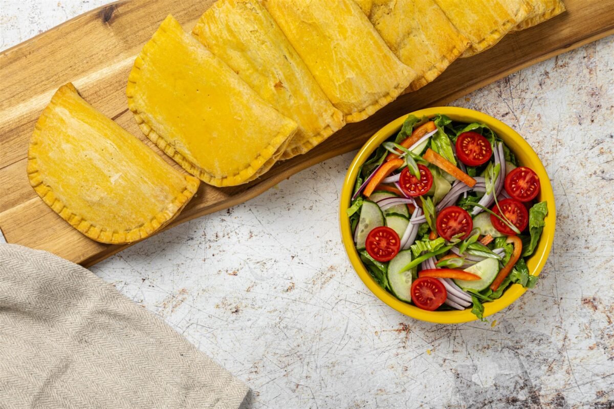 Overhead row of golden Jamaican patties on a light wooden board beside a yellow bowl of fresh salad
