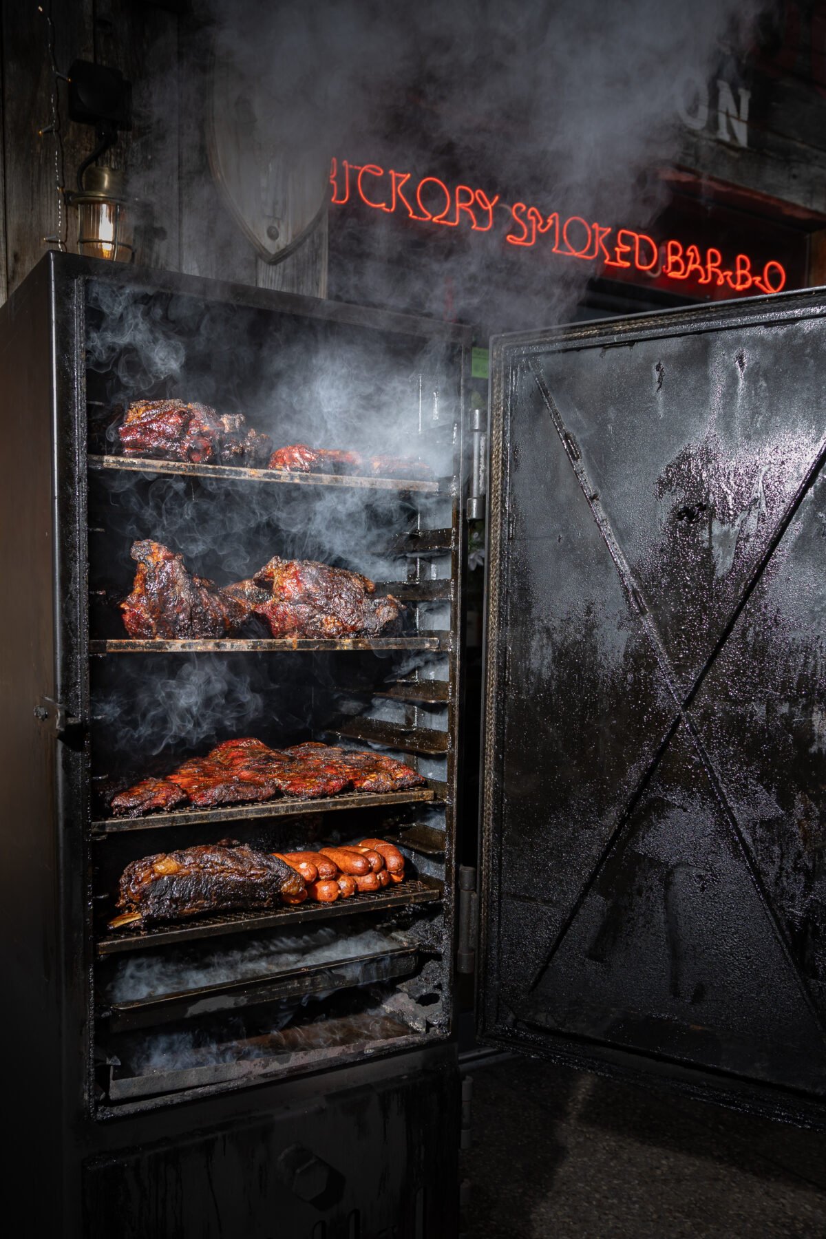 Smoker doors open with four loaded shelves, smoke rolling out under red neon signage