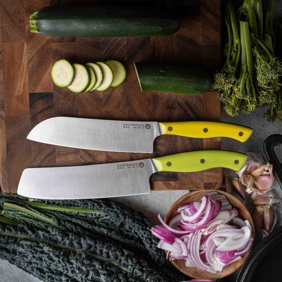Two yellow-handled kitchen knives on a wooden board with sliced courgette, red onion and greens