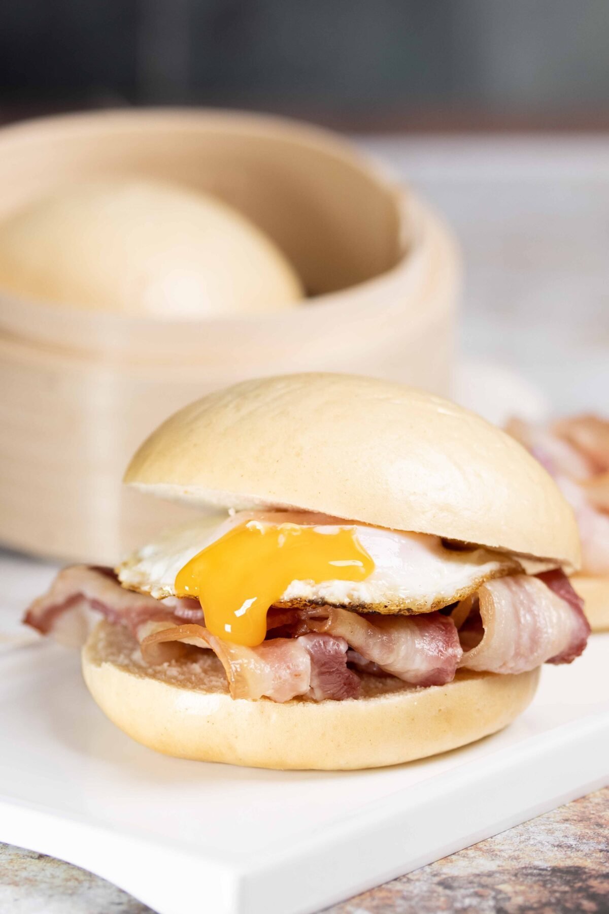 Bacon and fried egg bao bun on a white board with a bamboo steamer basket behind