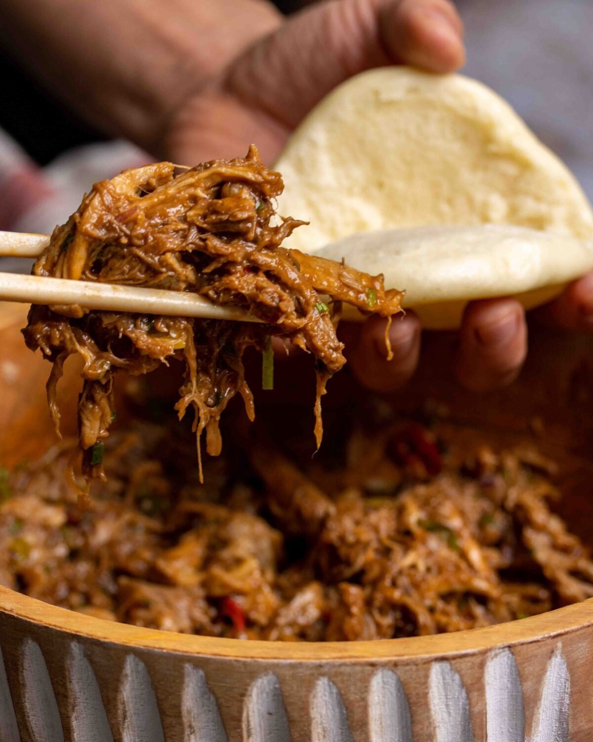 Chopsticks lifting pulled pork from a bowl beside a plain steamed bao bun