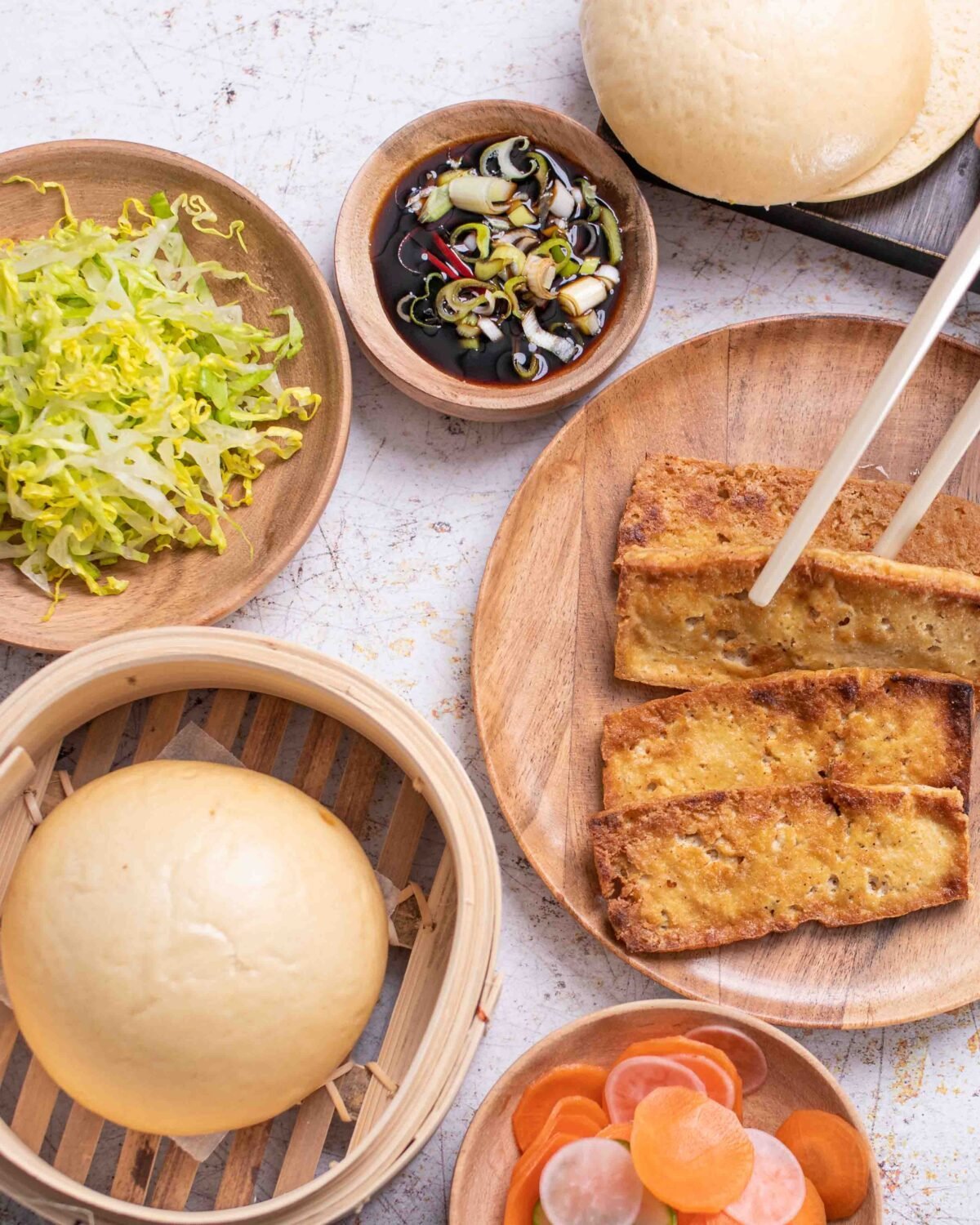 Overhead steamed bao buns, fried tofu, shredded lettuce and dipping sauces laid out on stone