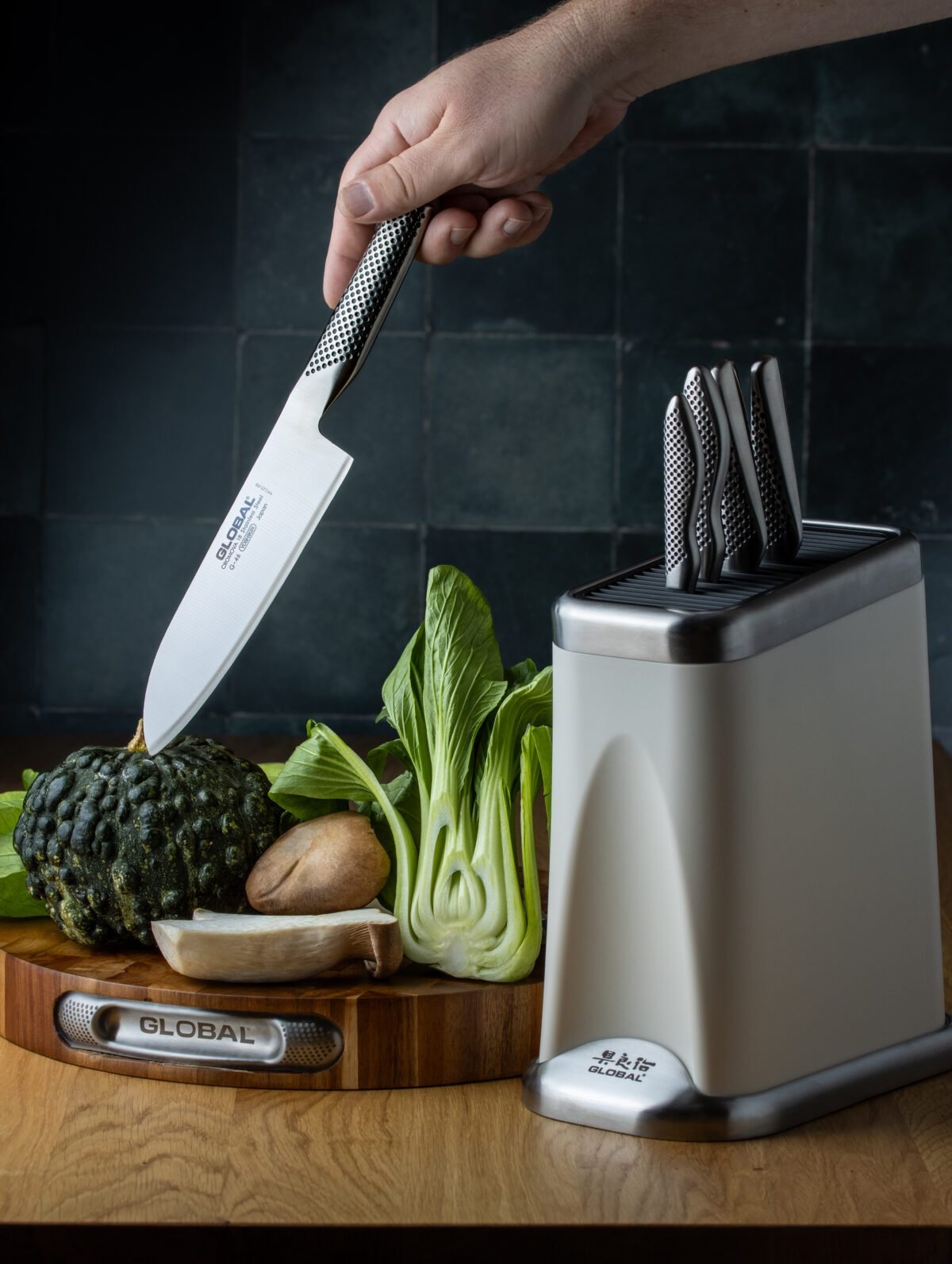 Hand lifting a Global santoku from a knife block, with broccoli, pak choi and potatoes on a board