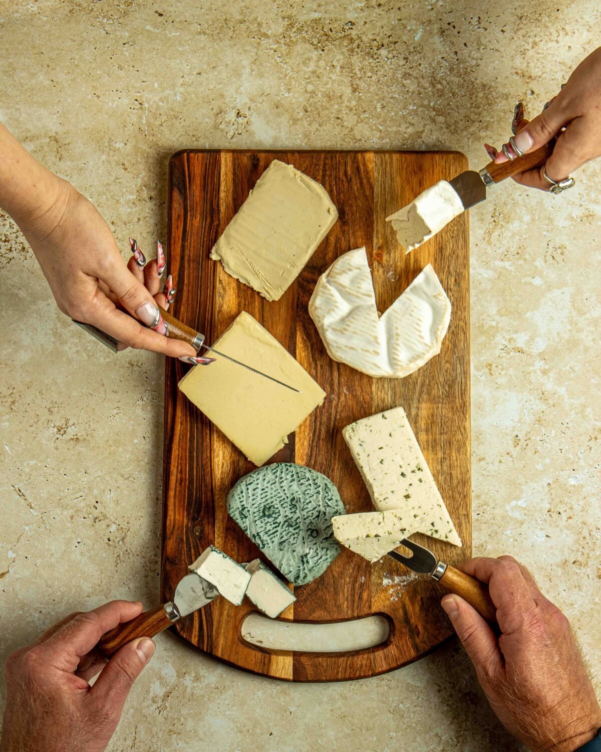 Four hands reaching across a cheese board with brie, blue cheese and hard cheeses