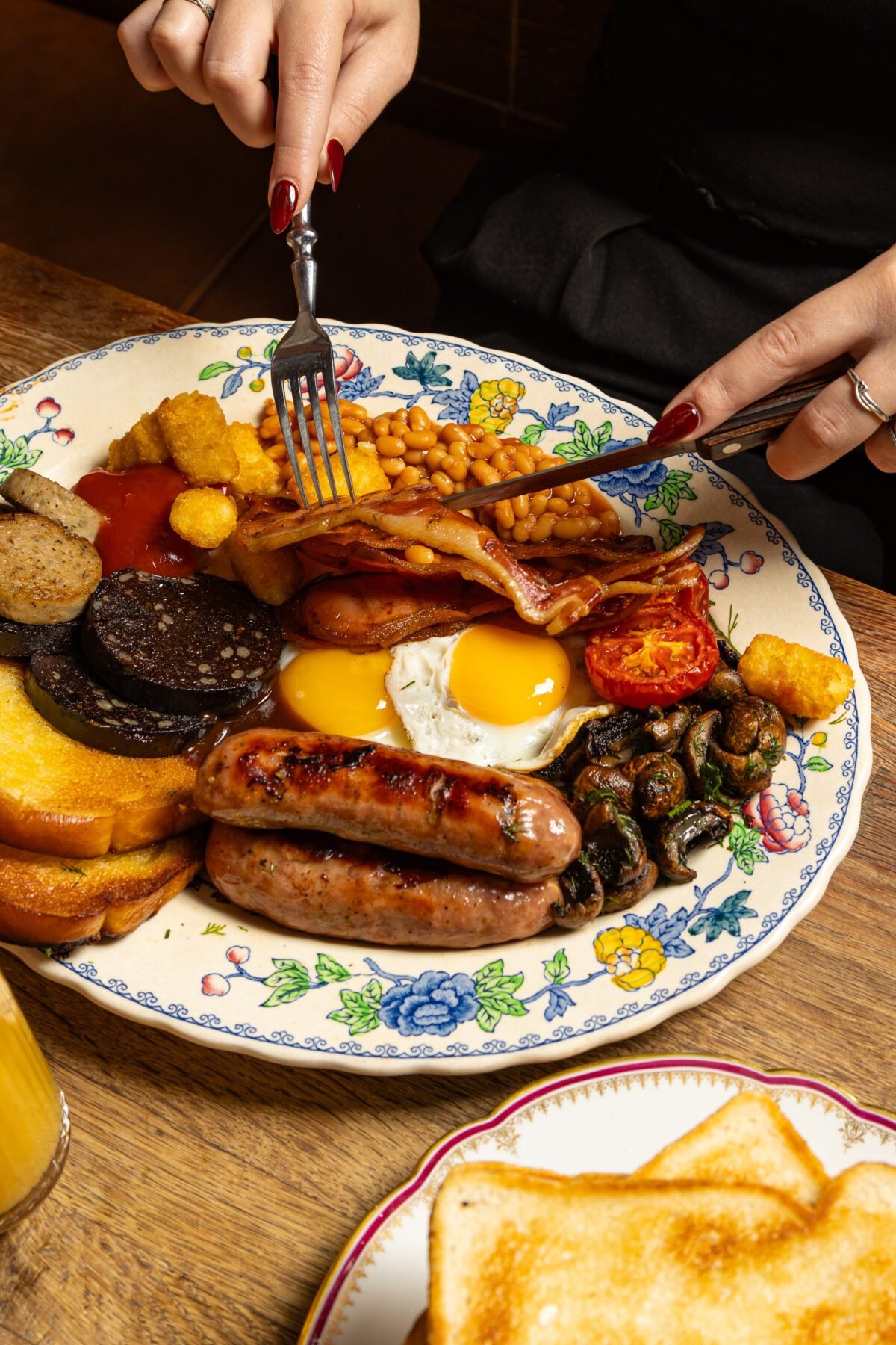 Full English breakfast on a floral plate being cut into with a knife and fork