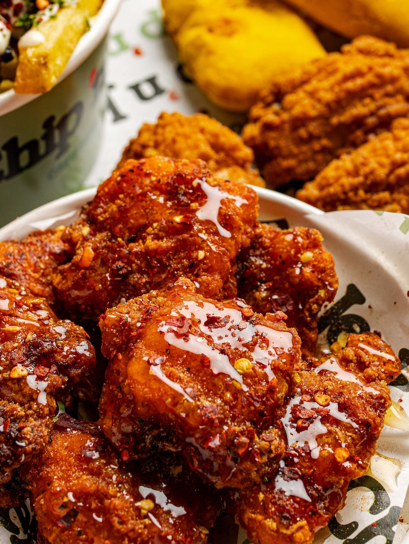 Close crop of sauced Caribbean fried chicken with a white drizzle and chilli flakes, with plantain and further chicken behind on branded paper, menu photography in Holloway, North London.
