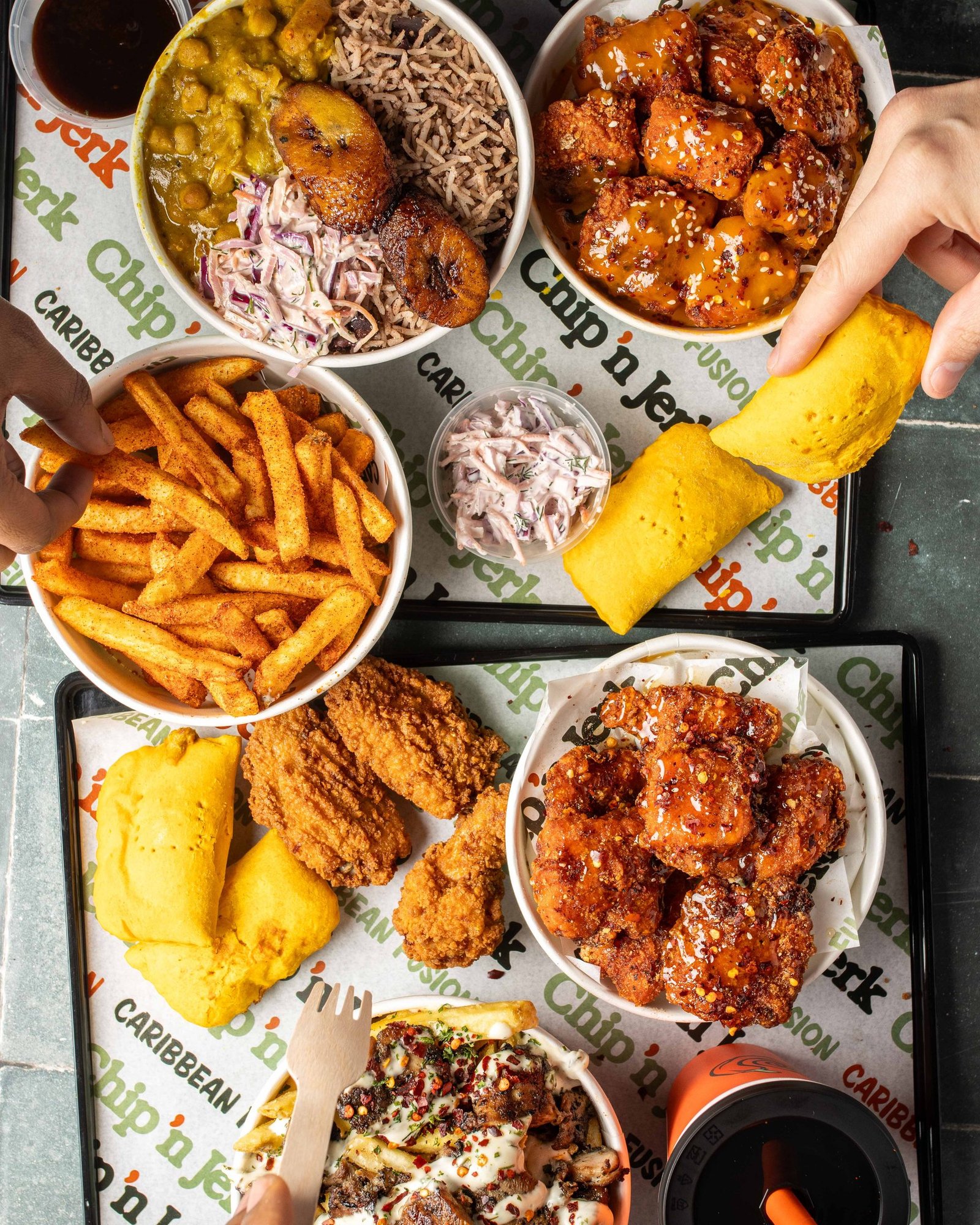Overhead of a full Chip 'n Jerk spread on branded trays, curry with rice and plantain, seasoned fries, wings, patties and slaw, with hands reaching in, lifestyle menu photography for a Caribbean QSR in Holloway.
