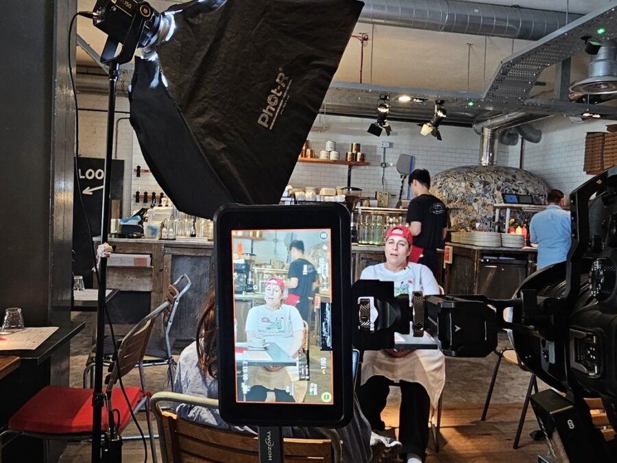 A chef being interviewed in front of the open kitchen at Franco Manca in Waterloo, lit with a softbox while the kitchen carries on behind, behind the scenes of a London restaurant film shoot.