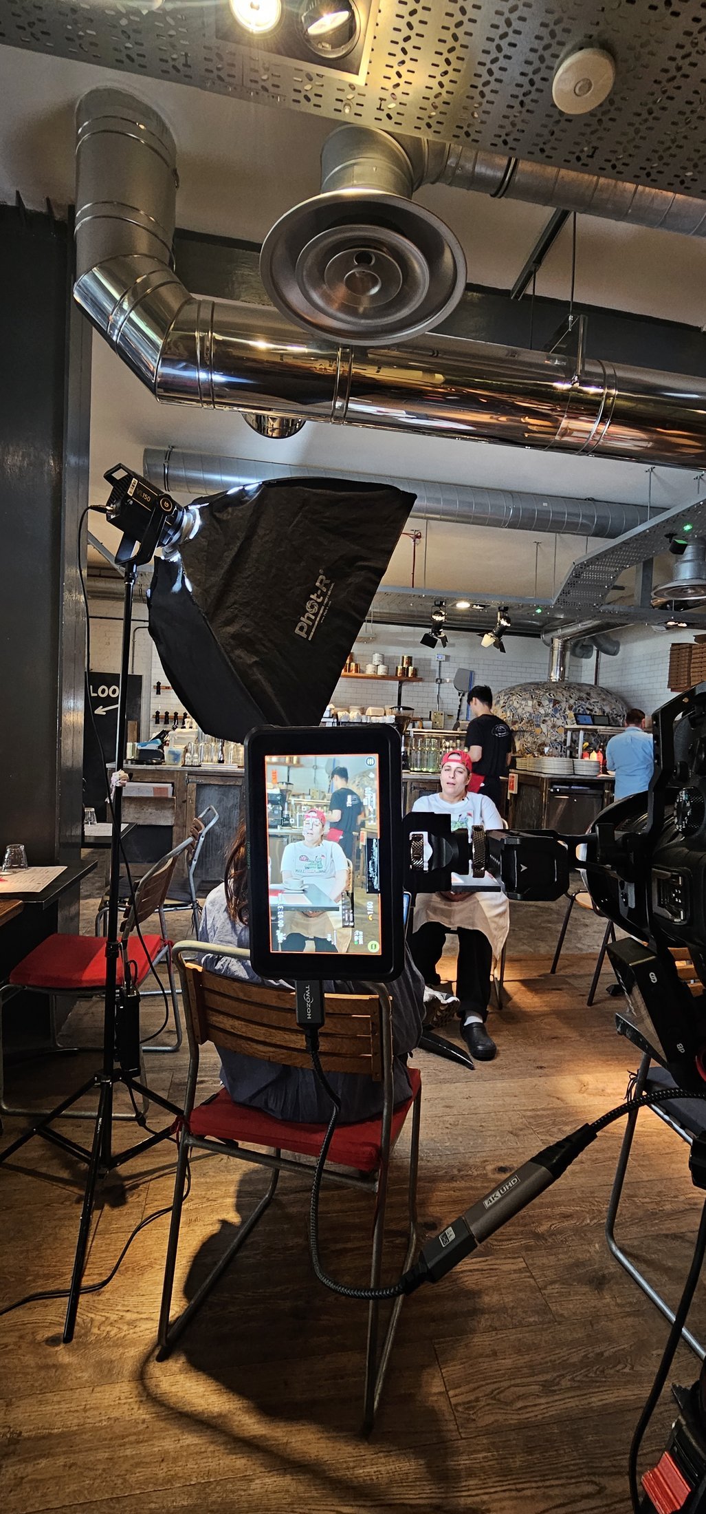 A chef being interviewed in front of the open kitchen at Franco Manca in Waterloo, lit with a softbox while the kitchen carries on behind, behind the scenes of a London restaurant film shoot.