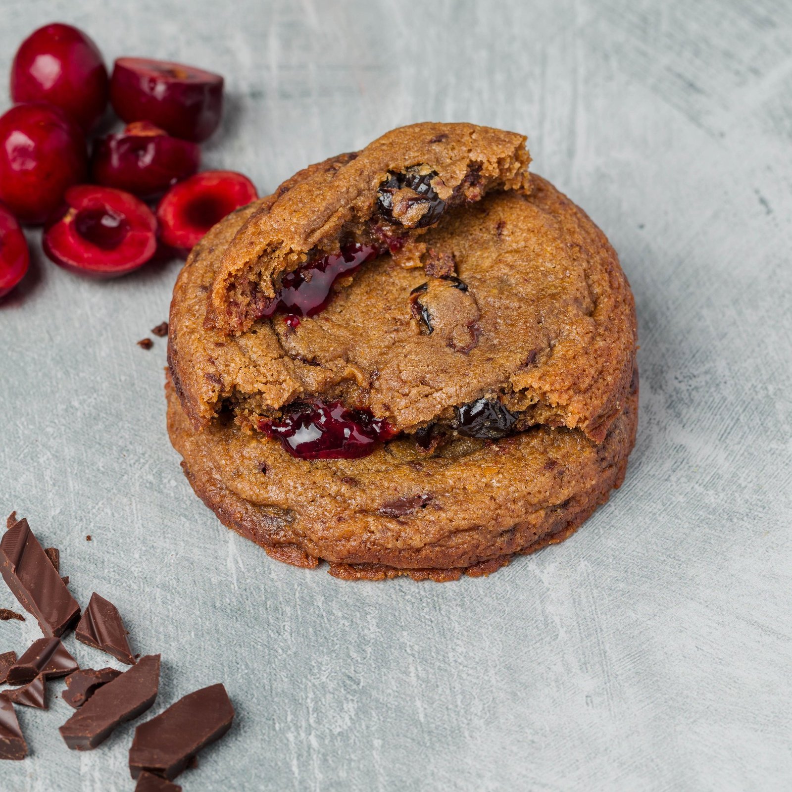 Cherry and dark chocolate cookie broken open to show a jammy cherry centre, styled with fresh cherries and dark chocolate, artisan cookie photography in Croydon.