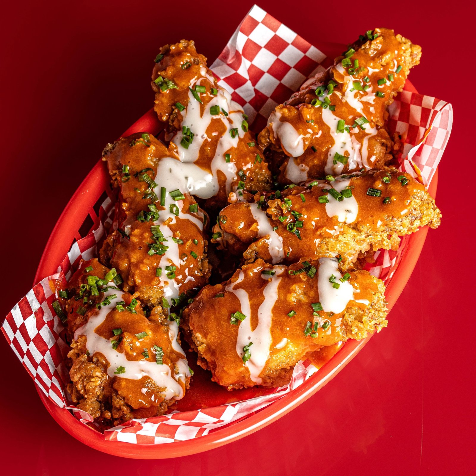 Chicken wings in a red basket on checked paper, glazed in hot sauce with a ranch drizzle and chopped chives, delivery menu photography for a chicken shop in Wood Green, North London.