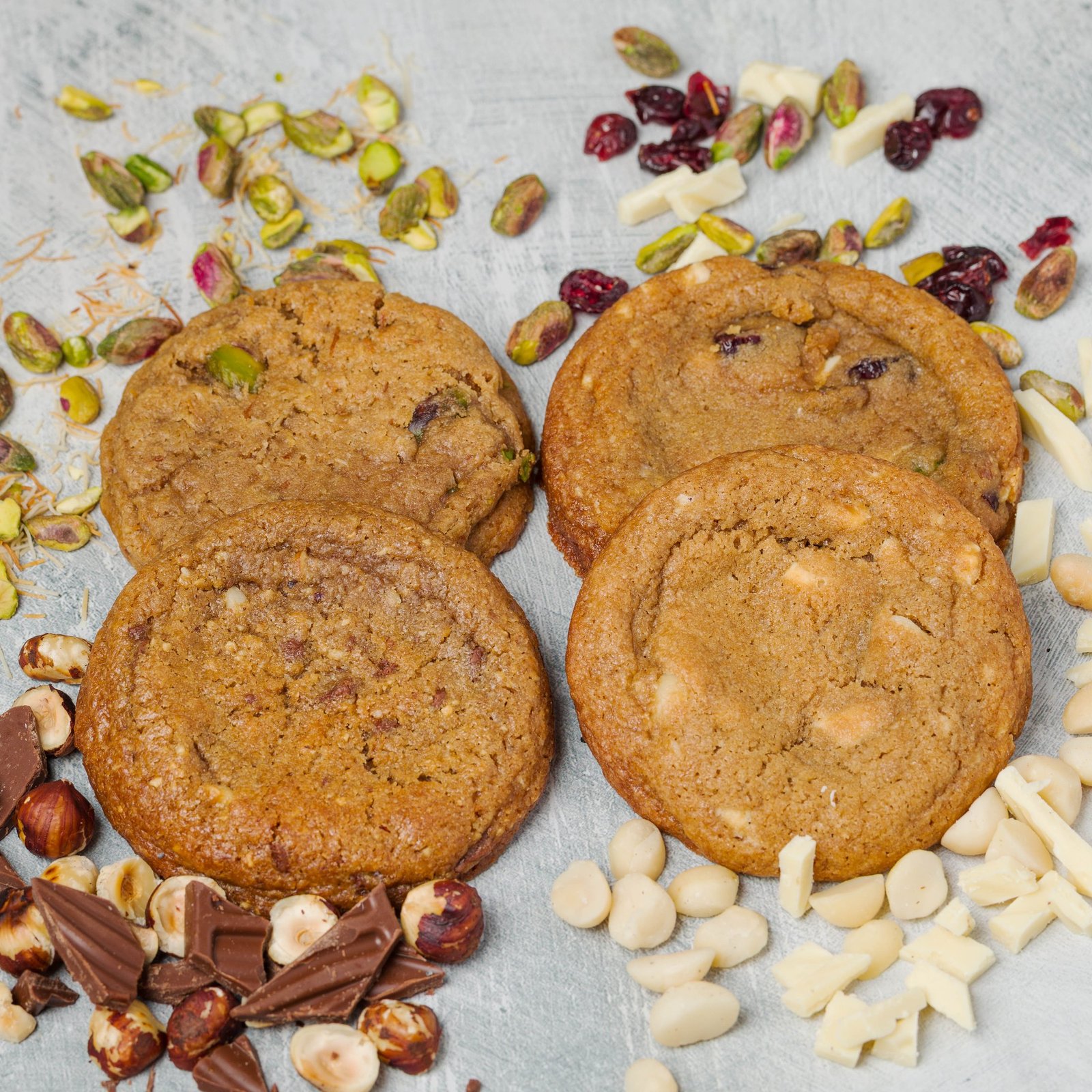 Overhead flat lay of four cookie flavours with their own ingredients scattered around them, pistachio, cranberry, hazelnut, white chocolate and macadamia, food styling for a Croydon cookie brand.