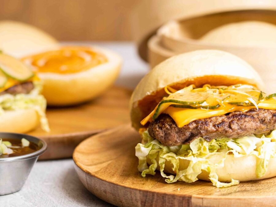 A bao bun used as a burger bun, filled with a beef patty, melted cheese and shredded lettuce on a wooden board, with a second sauced bao and a dipping pot behind, food photography and food styling for a bao producer in Acton, London.