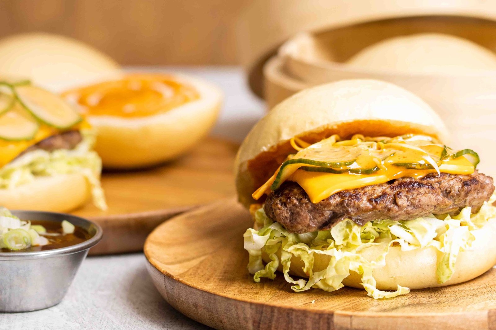 A bao bun used as a burger bun, filled with a beef patty, melted cheese and shredded lettuce on a wooden board, with a second sauced bao and a dipping pot behind, food photography and food styling for a bao producer in Acton, London.