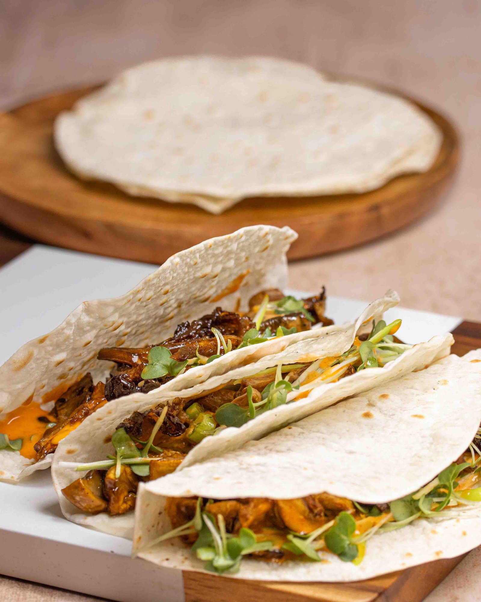 Three Chinese pancakes folded around shredded pork with sauce and microgreens, a plain pancake on a wooden board behind, food photography for a pancake manufacturer in London.