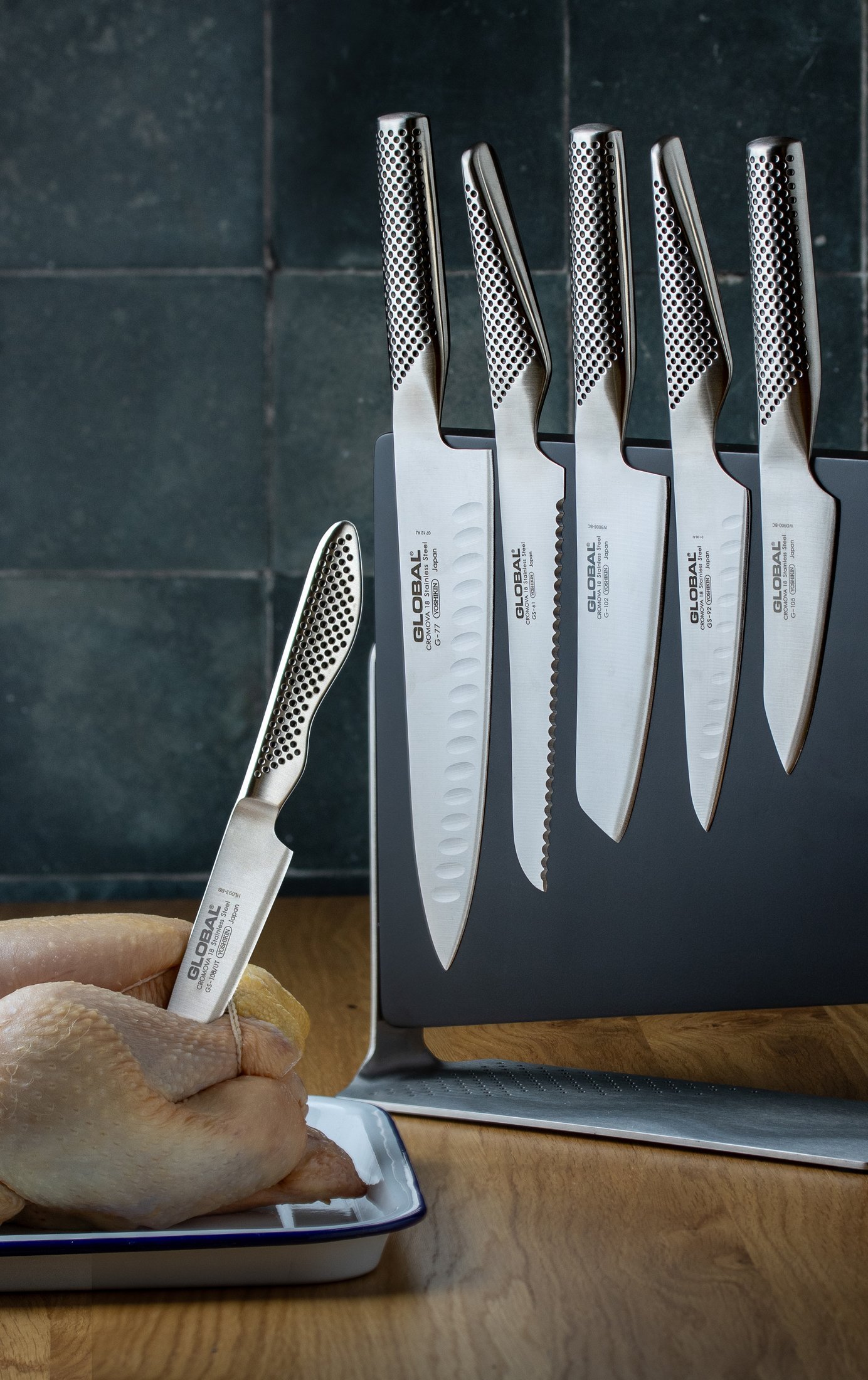 A Global magnetic knife rack holding five premium Japanese knives with a small Global knife standing in a whole chicken in front, food photography for the Air knife range.