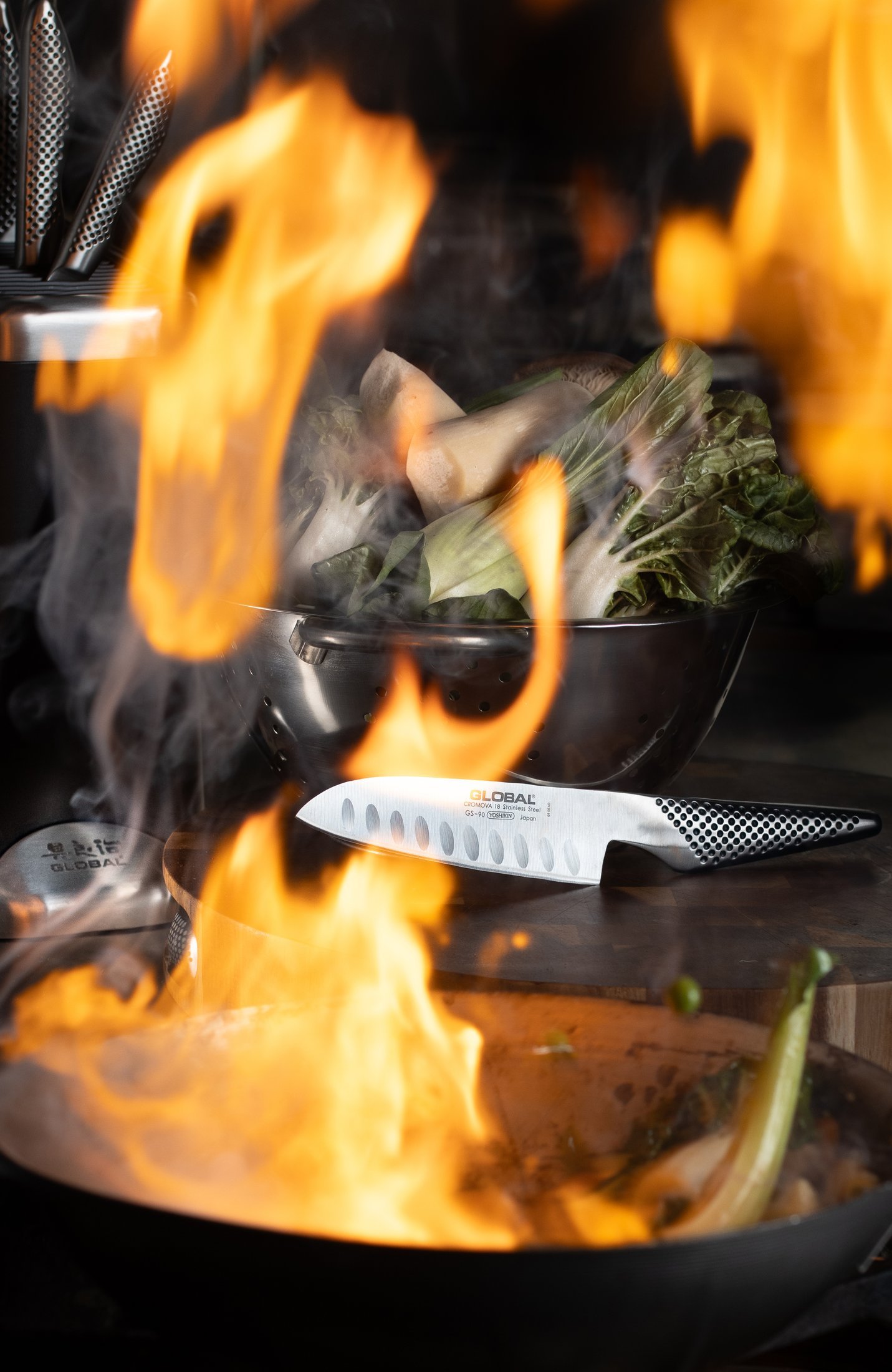 Flames rising around a wok and a colander of bok choy and king oyster mushrooms, a Global fluted santoku resting on the board, food photography for the Fire knife range.