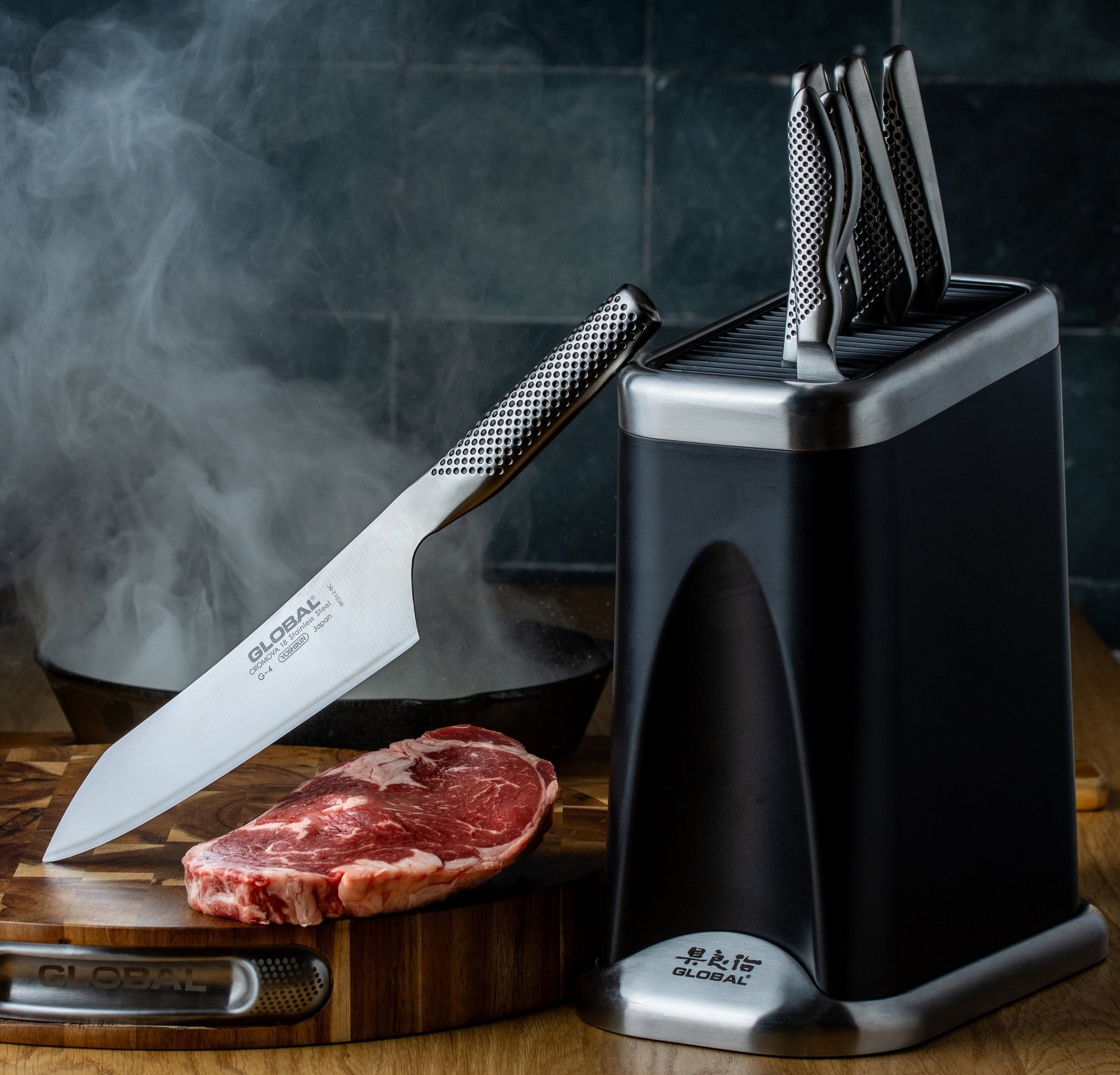 A ribeye steak on a wooden board with a Global knife leaning against the knife block and steam rising from a hot pan behind, food photography for a premium kitchen knife brand.