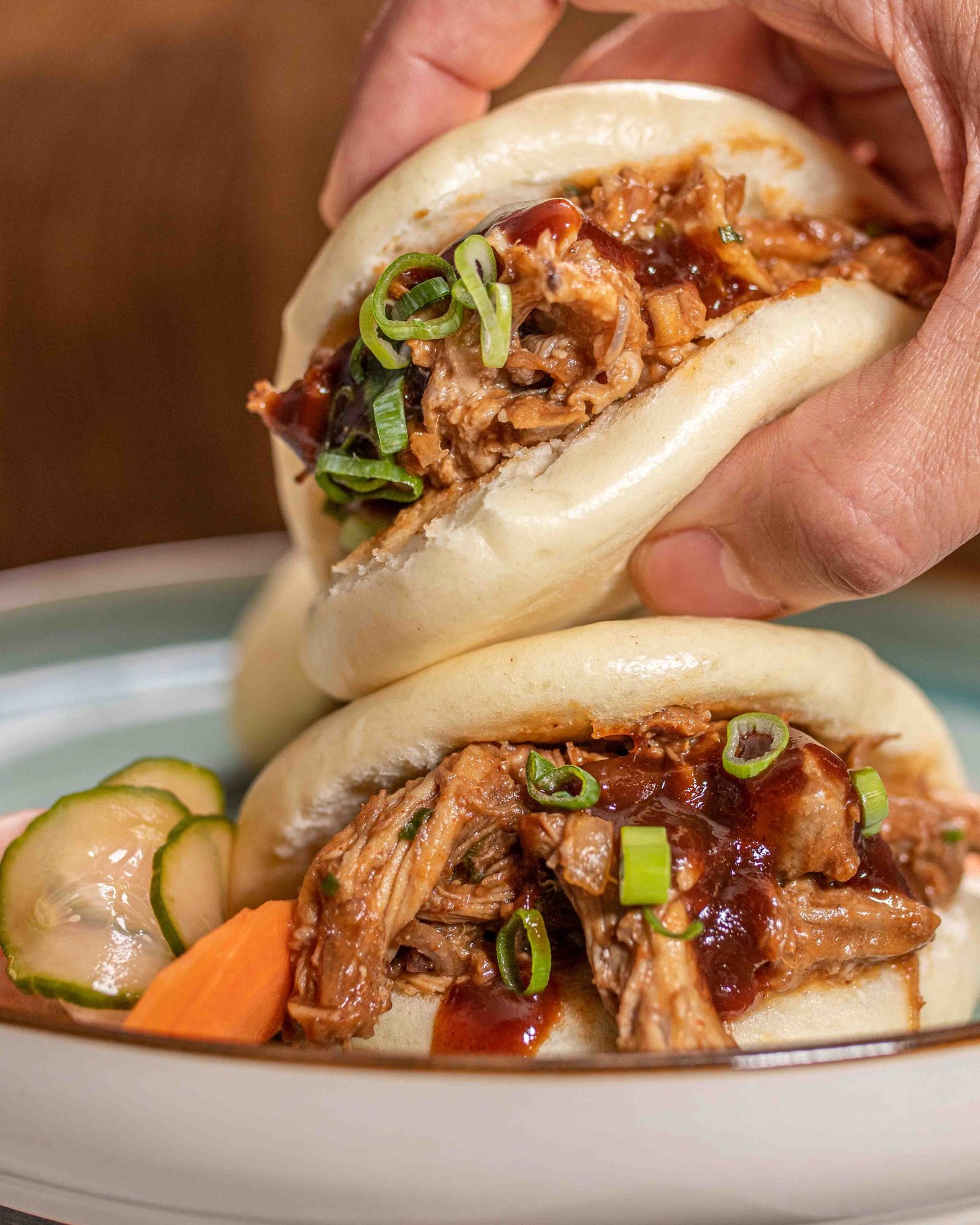 A hand lifting a pulled pork bao topped with spring onion and sauce above a plate holding a second bao with pickles, lifestyle food photography for a food manufacturer in London.