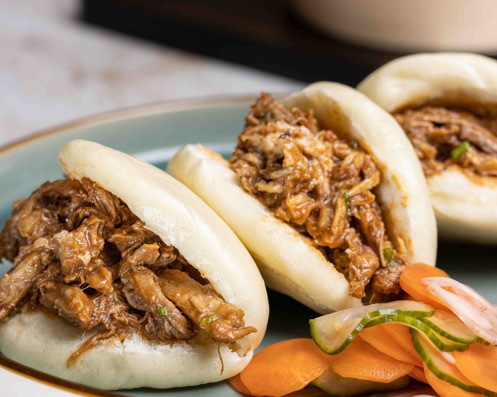 Two Hirata bao filled with pulled pork, spring onion and sauce on a pale blue plate with pickled carrot and cucumber, lifestyle food photography for a food manufacturer.