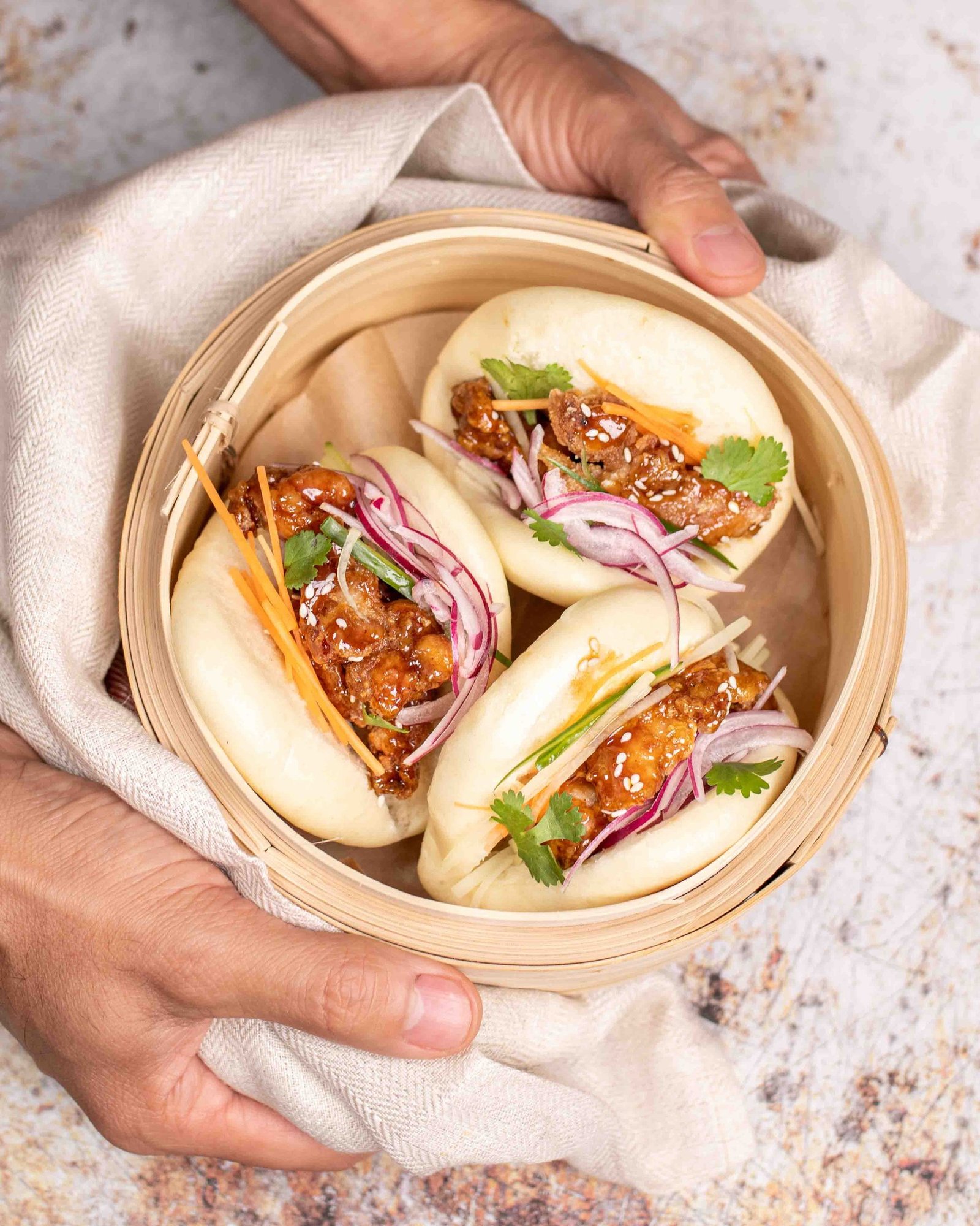 Two hands holding a cloth-lined bamboo steamer with two bao filled with teriyaki chicken, red onion and coriander, lifestyle food photography for a bao producer.