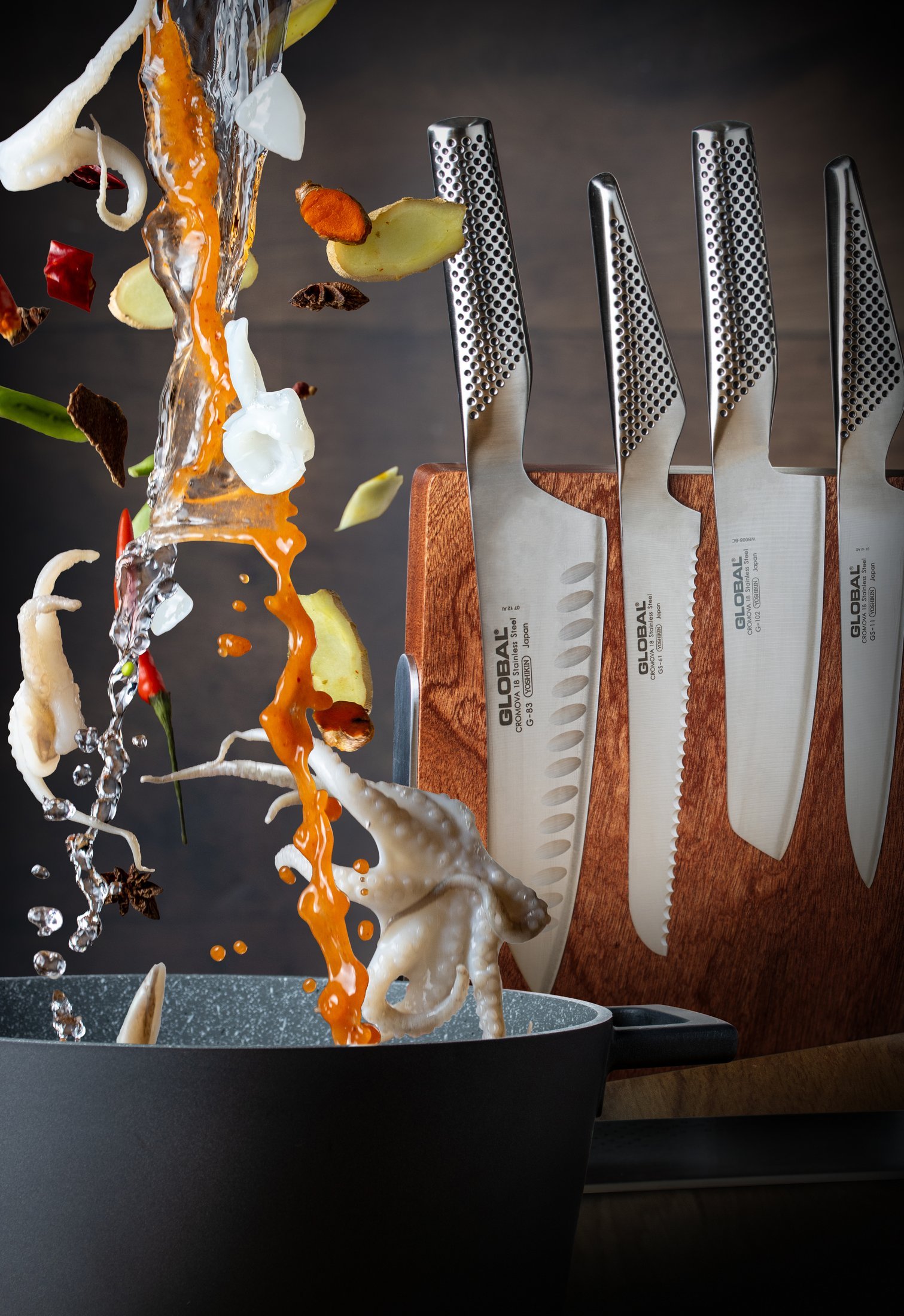 Squid, calamari rings, red chilli, sliced ginger, turmeric and star anise suspended in mid-air in a stream of orange broth falling into a dark pan, beside a Global wooden magnetic board holding four premium Japanese knives, food styling and photography for the Water knife range.
