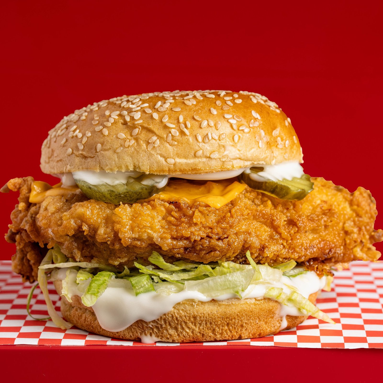 A fried chicken burger in a sesame bun with cheese, pickles, shredded lettuce and mayo, photographed side on against red, delivery menu photography in Wood Green, North London.