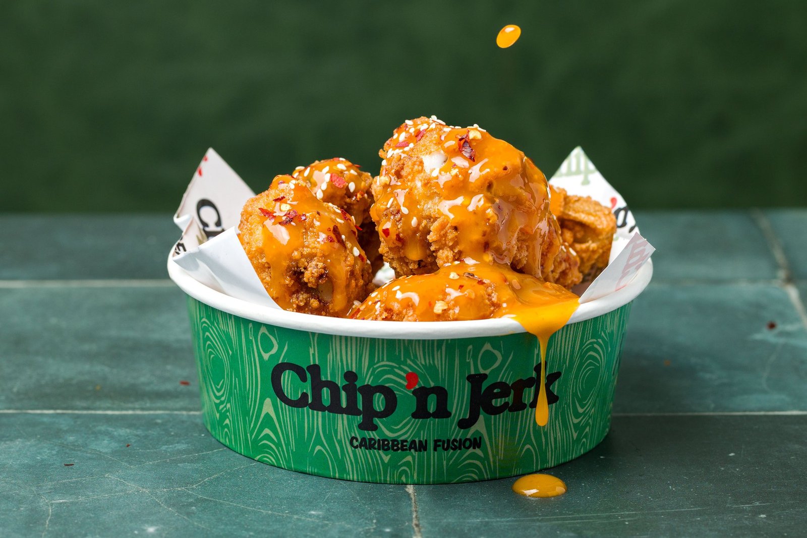 Fried chicken in a branded tub finished with a golden mango-coloured sauce, a droplet caught in mid-air above the bowl, delivery menu photography for a Caribbean fusion QSR in Holloway.