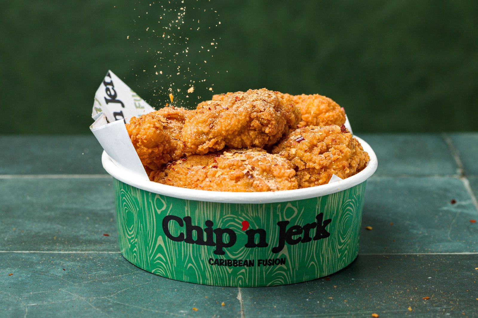 Fried chicken in a branded Chip 'n Jerk tub with seasoning falling through the frame, food styling and menu photography for a Caribbean QSR in Holloway, North London.